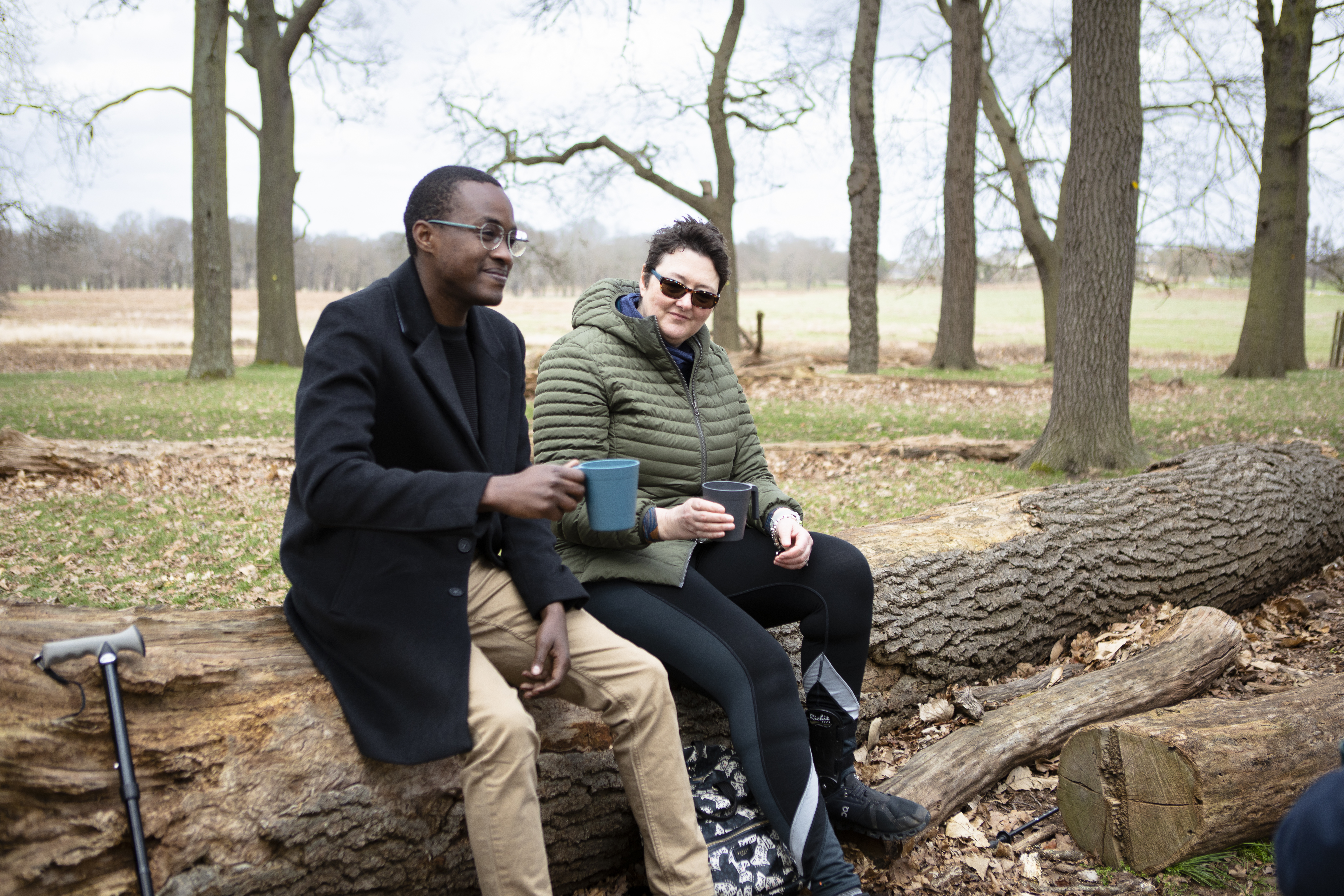 Veterans Denis and Debbie are sitting on a log in a green field, chatting over a hot drink
